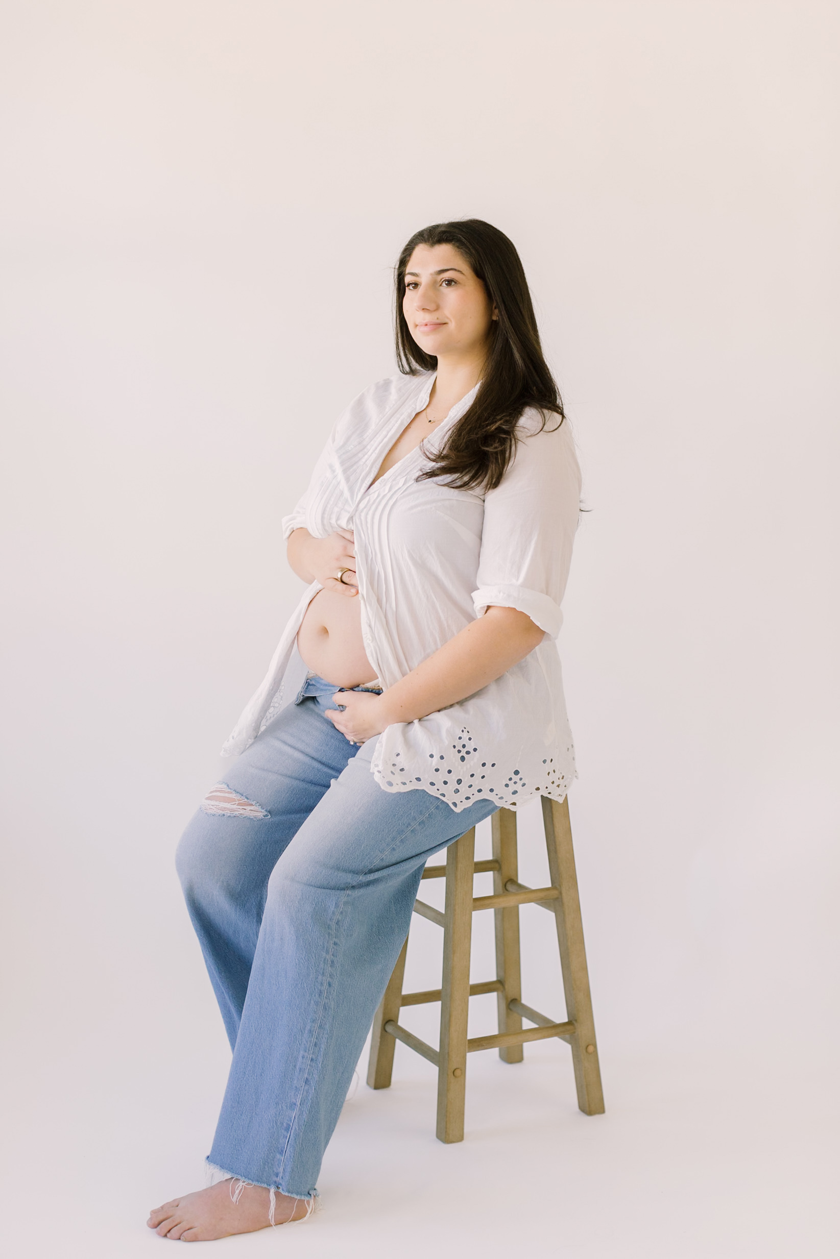 A pregnant lady sits on a stool as she smiles and stares into the distance of a white studio while holding her bump.