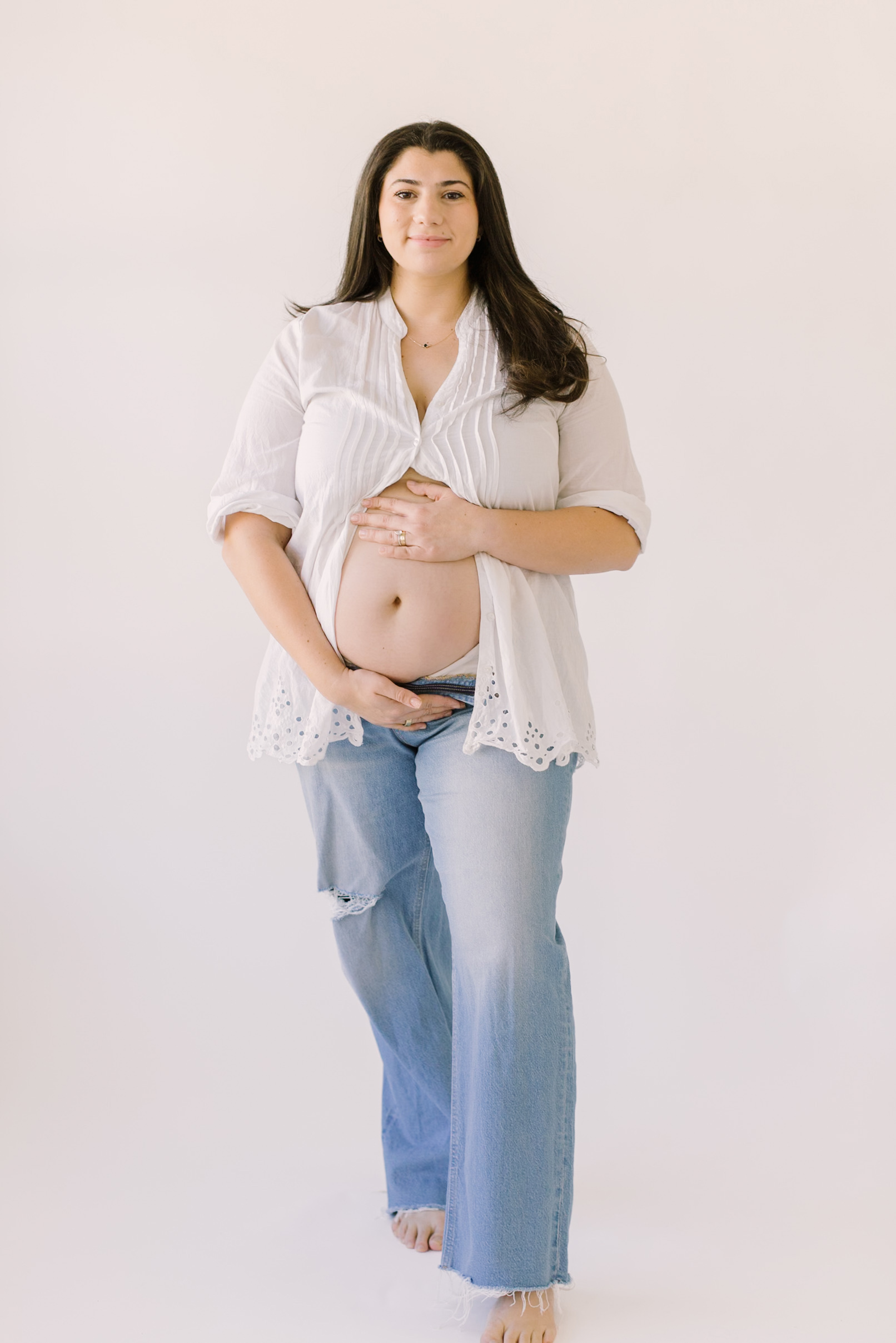 A soon to be mother holds her bump as she smiles at the camera in a white backgrounded studio wearing jeans and a white button up that shows her bump.