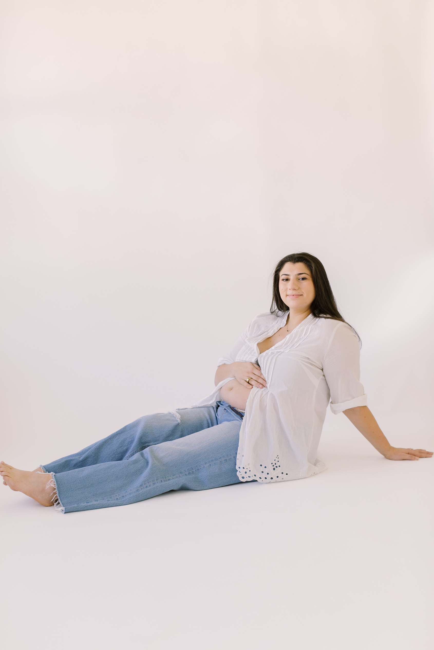 A mother sits on the floor of a white studio with her legs out as she holds one hand on her bump and smiles after a session of prenatal yoga in raleigh.
