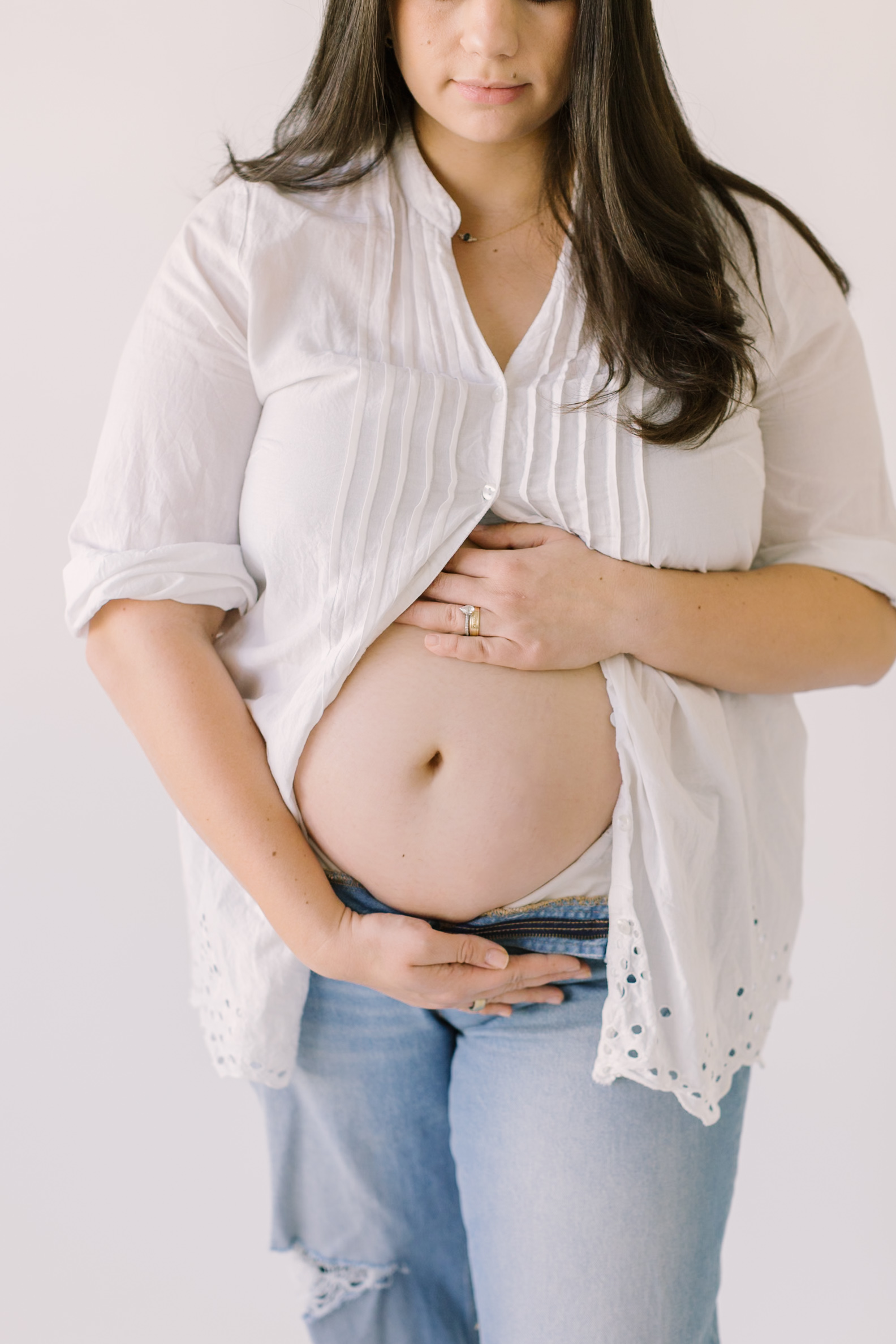 A mom wearing a white button up and jeans looks down and holds her exposed bump after prenatal yoga in raleigh.