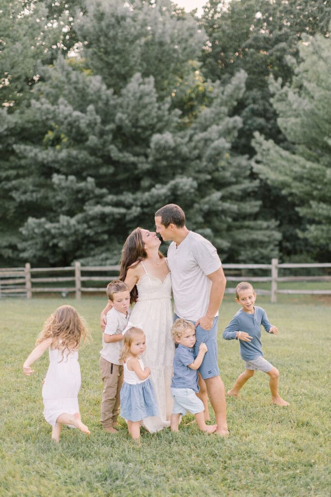Family of eight during an outdoor Raleigh family photography session, captured in a playful moment with barefoot kids running and parents sharing a kiss.