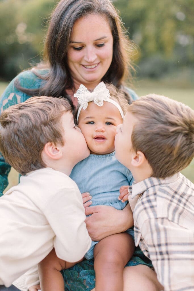 Mother holding baby girl while two brothers kiss her cheeks during a Raleigh family photography session, highlighting sibling connection and joy.