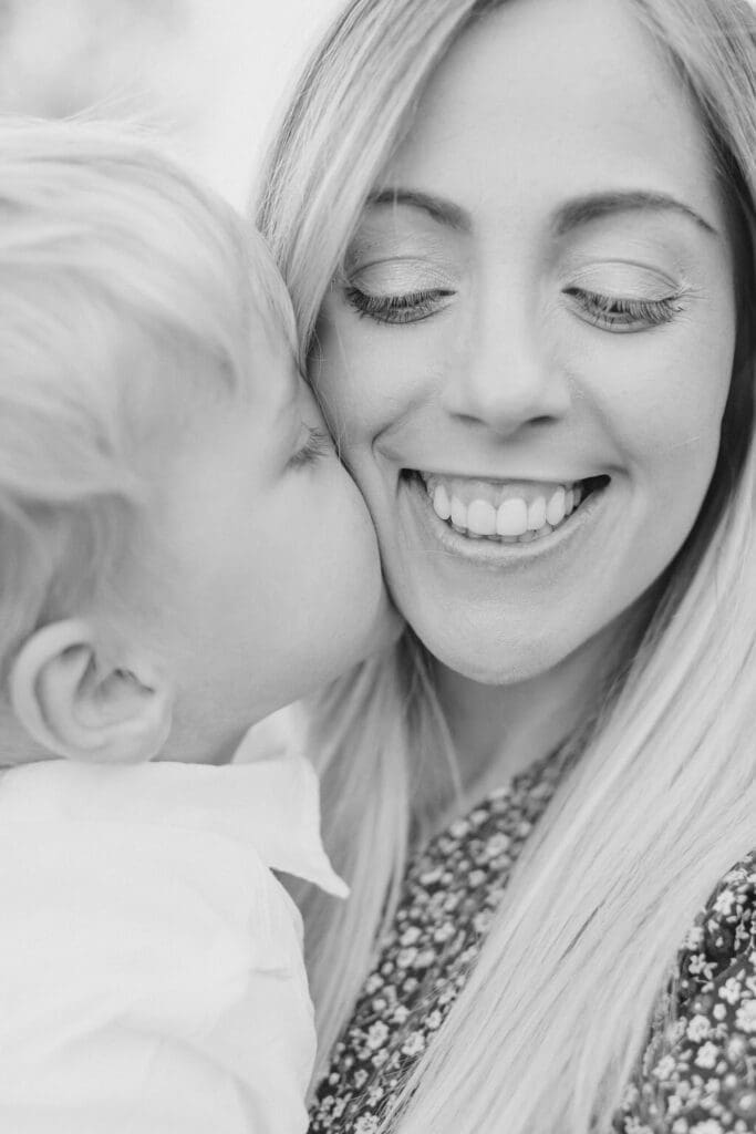 Close-up black and white portrait of a mother smiling as her young son kisses her cheek, captured during a heartfelt Raleigh family photo session.