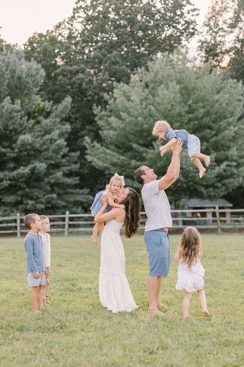 Large family playing outdoors during a Raleigh family photo session, with parents lifting toddlers and siblings watching joyfully in a natural, open field.