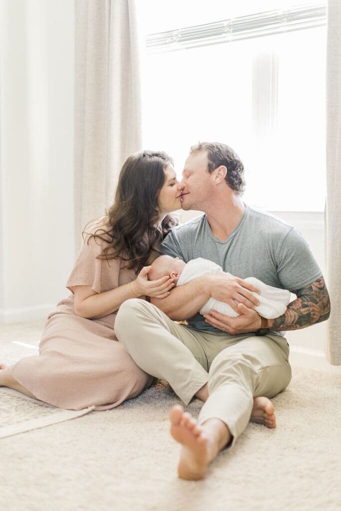 Parents sitting on the floor in natural light during a Raleigh newborn photography session, kissing while holding their swaddled baby.