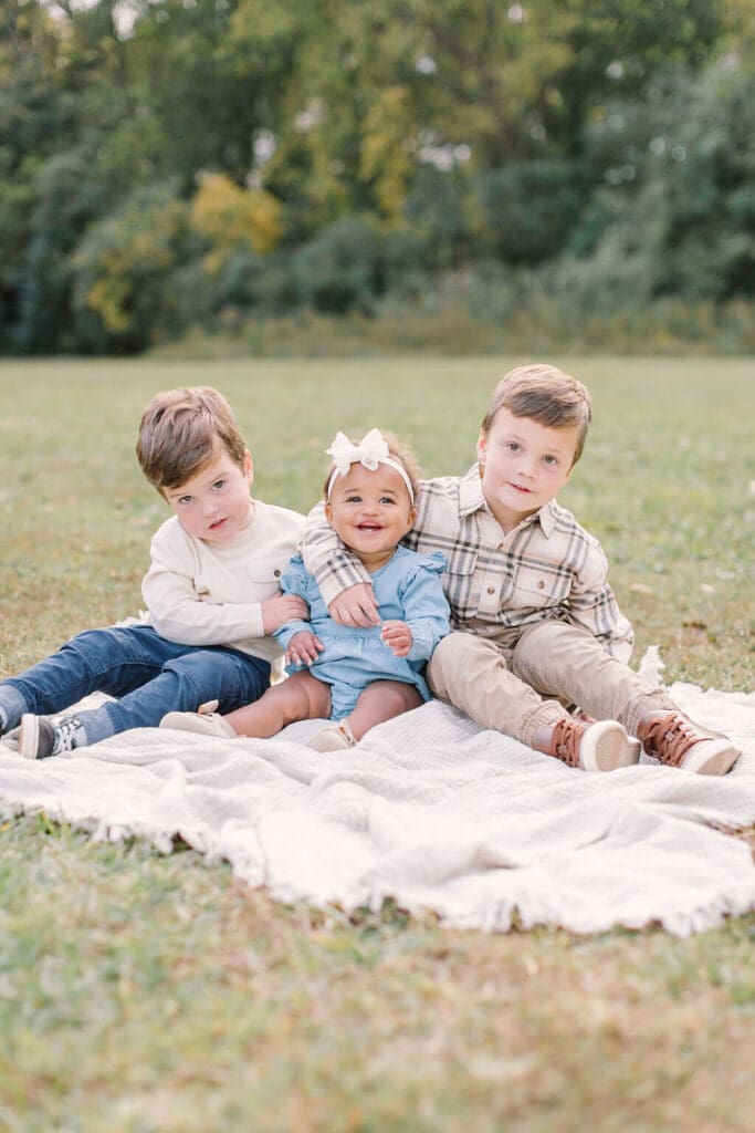 Three young siblings sitting on a blanket during a Raleigh NC family photography session, with the baby girl smiling between her two big brothers.