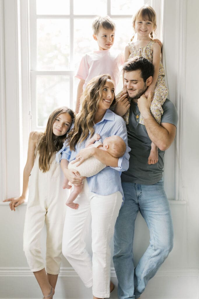 Family of six standing by a large window during a lifestyle newborn session in Raleigh, smiling while mom holds the new baby in soft natural light.