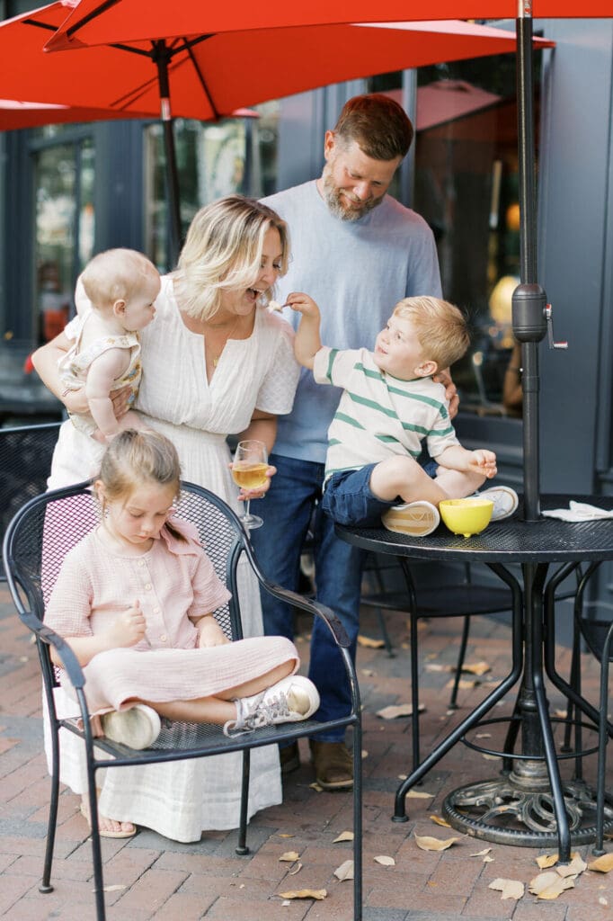 Candid lifestyle family session in downtown Raleigh, with parents and three children sharing a snack at an outdoor café under bright red umbrellas.