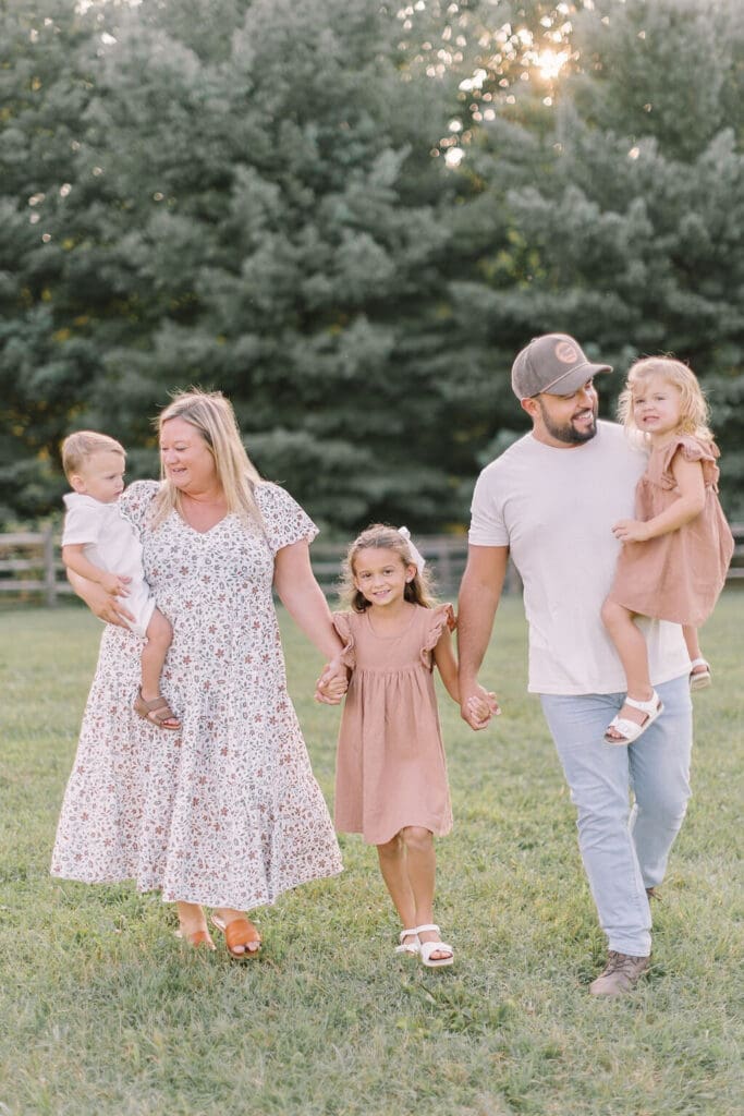 Family of five walking together during a Raleigh NC family photography session, smiling and holding hands as the sun sets behind them in the trees.