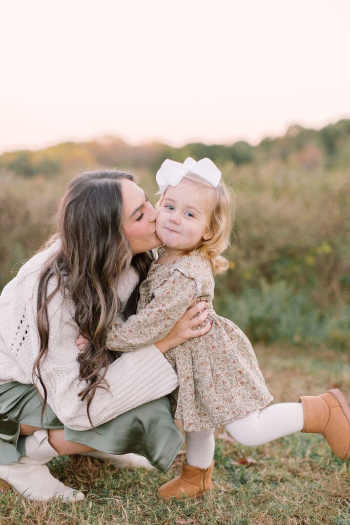 Mother kissing her toddler daughter on the cheek during a Raleigh family session, with soft light and cozy fall outfits in a warm outdoor setting.