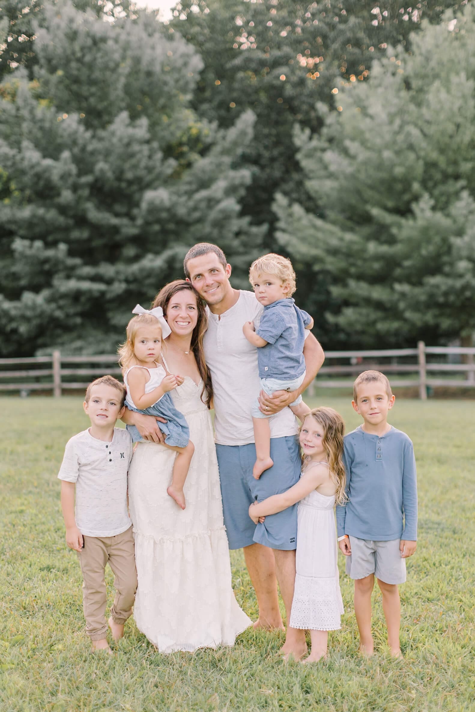 A mom and dad stand with their five young children in a meadow at sunset as they all smile