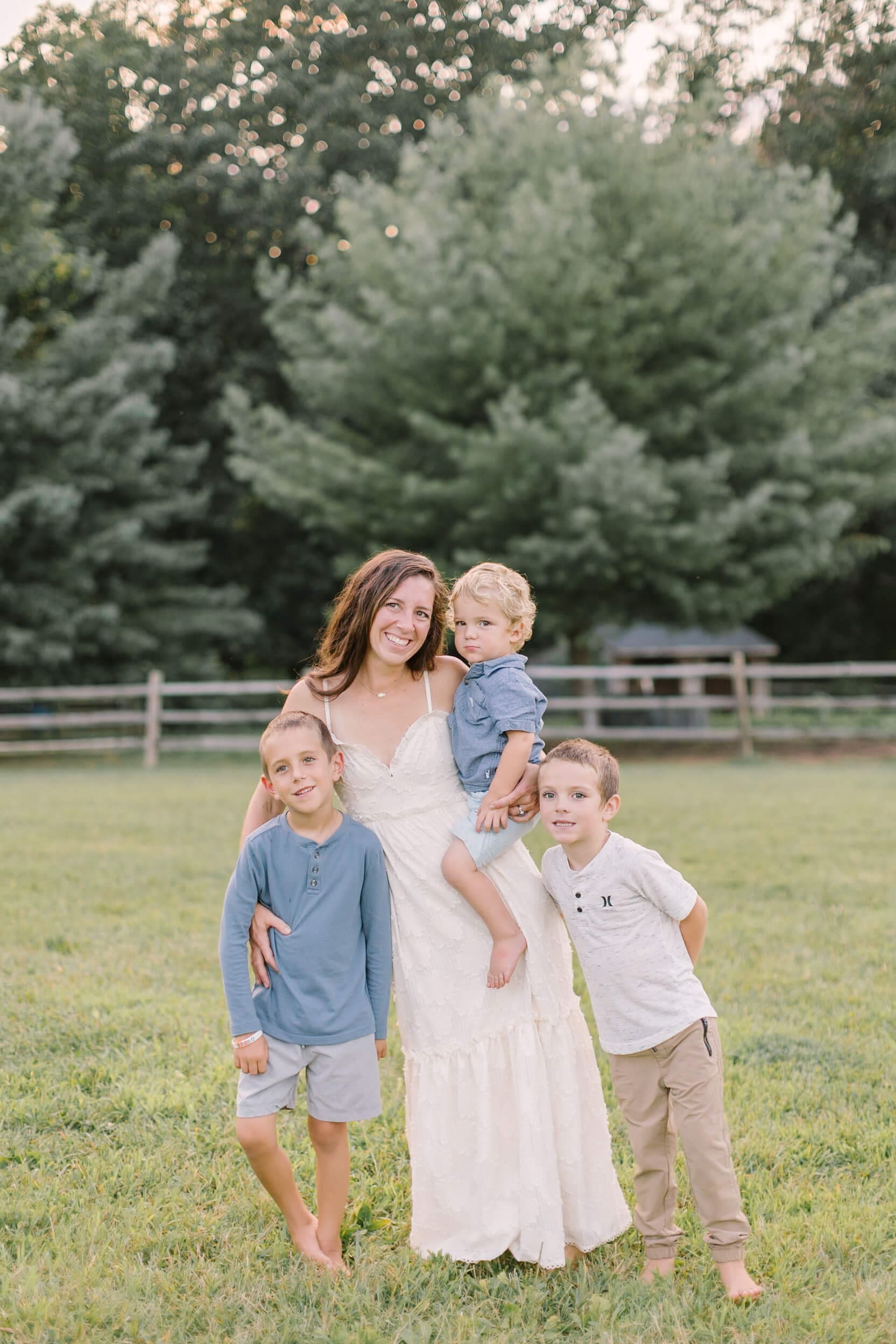 A smiling mom stands with her three sons in a pasture at sunset in a white dress after some apple picking in raleigh