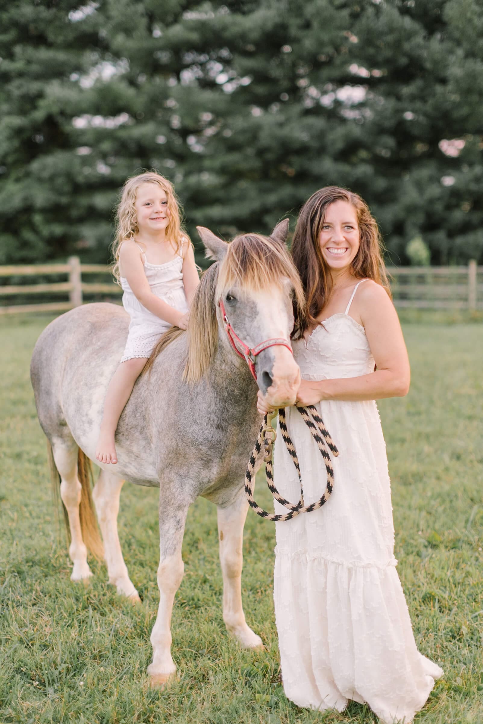 A happy mom in a white dress helps her daughter ride bareback on a pony in a pasture after some apple picking in raleigh