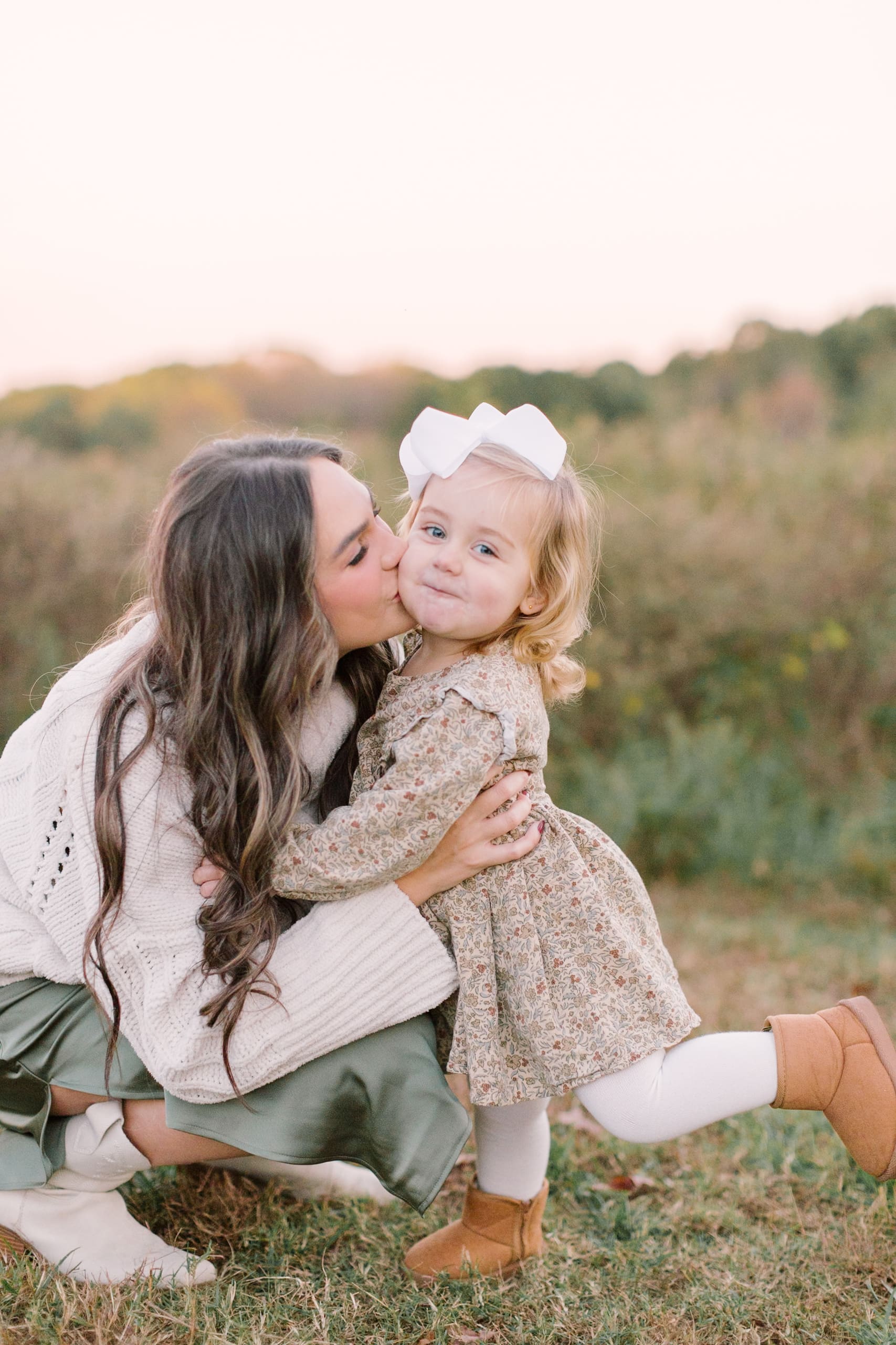A happy toddler girl in a green dress is kissed by mom in a park trail at sunset