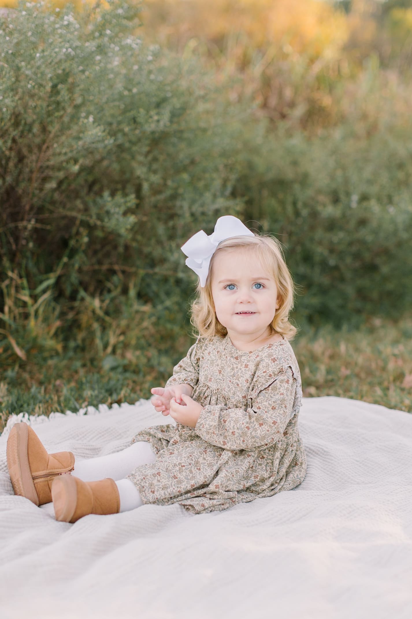 A toddler girl in a green dress and large white bow sits on a picnic blanket at sunset after visiting boutiques in raleigh
