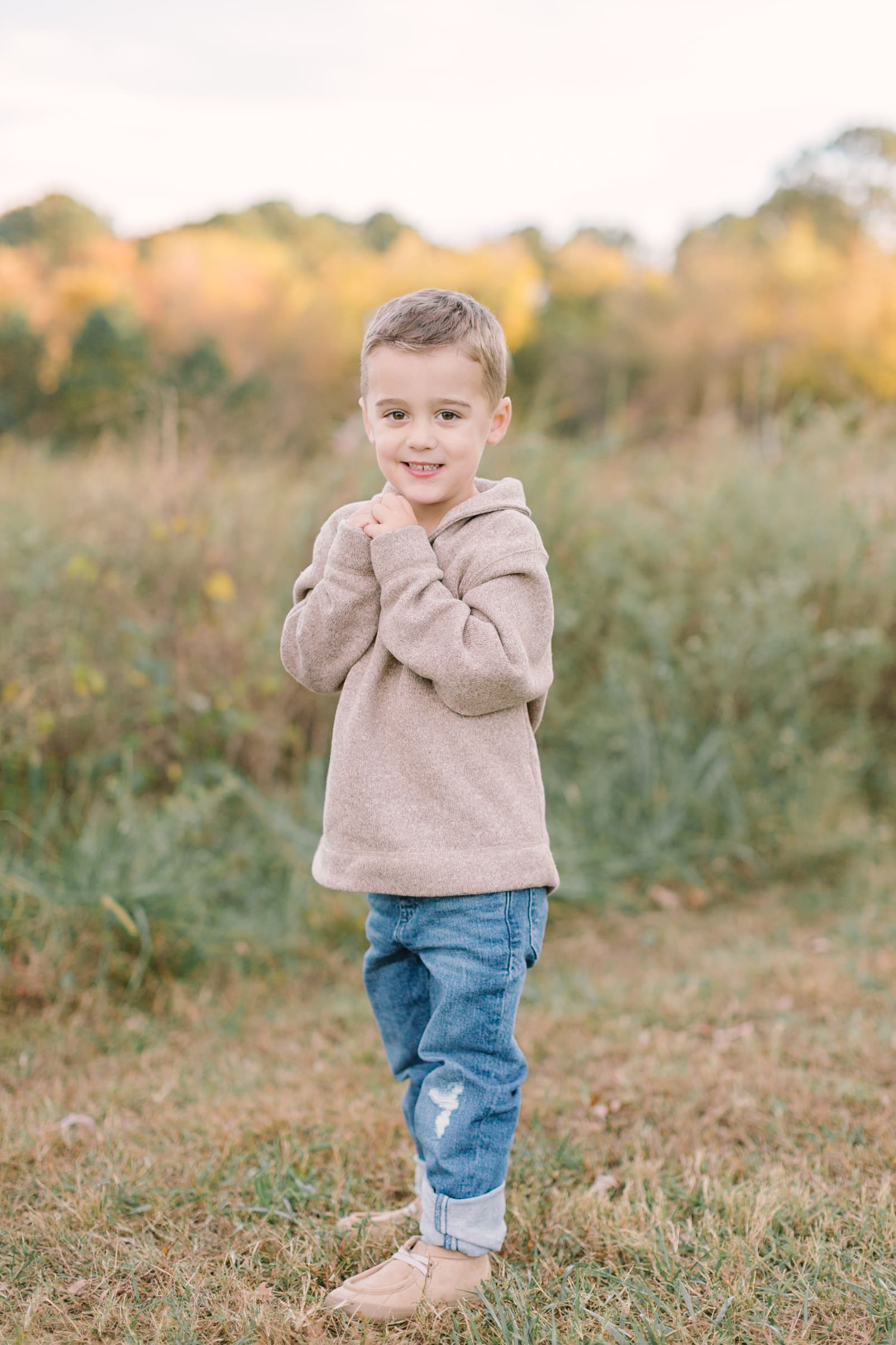A smiling toddler boy in a tan sweater and jeans stands in a park trail at sunset after visiting boutiques in raleigh