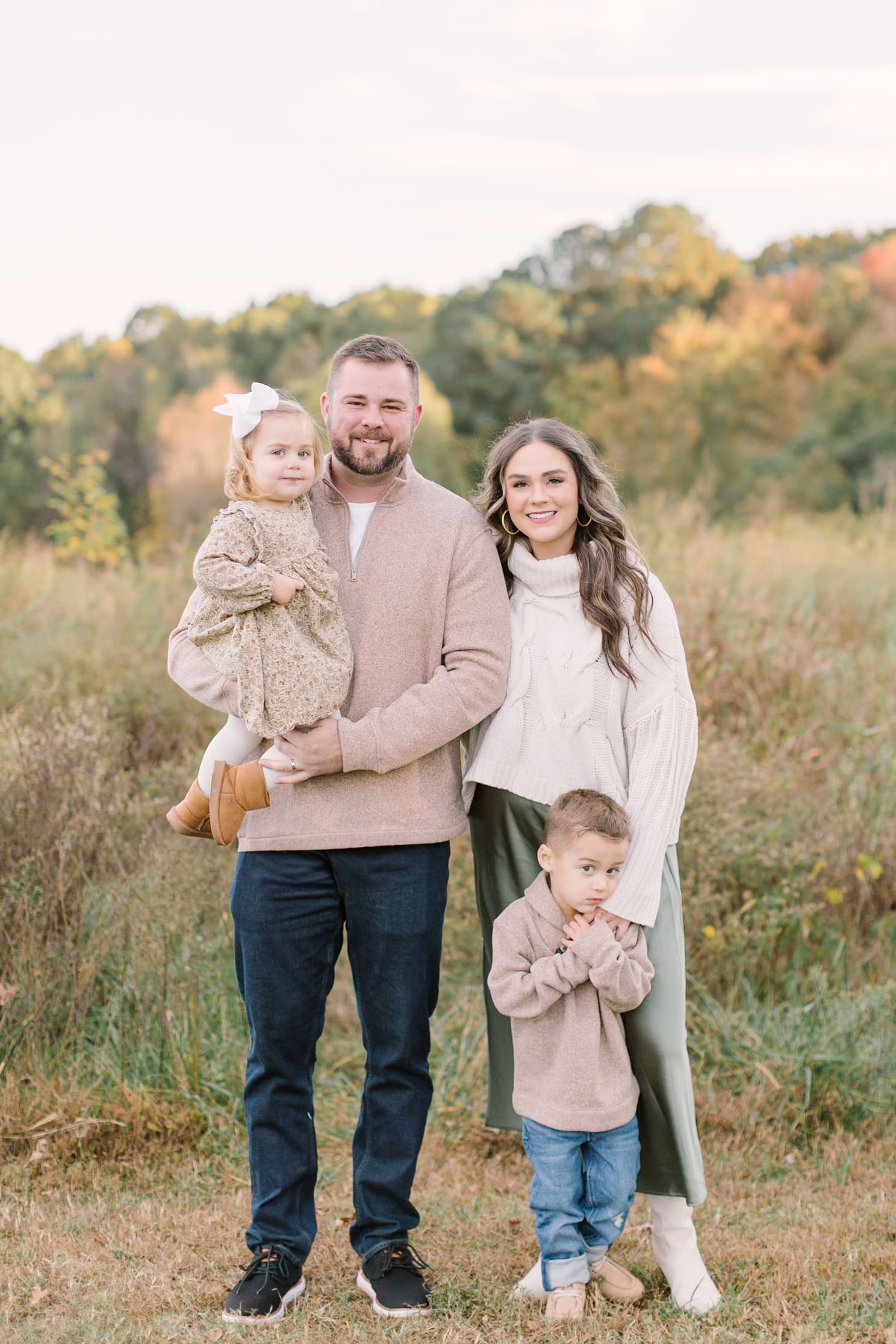 A mom and dad stand in a park trail through tall grass at sunset in fall with their toddler son and daughter after visiting boutiques in raleigh