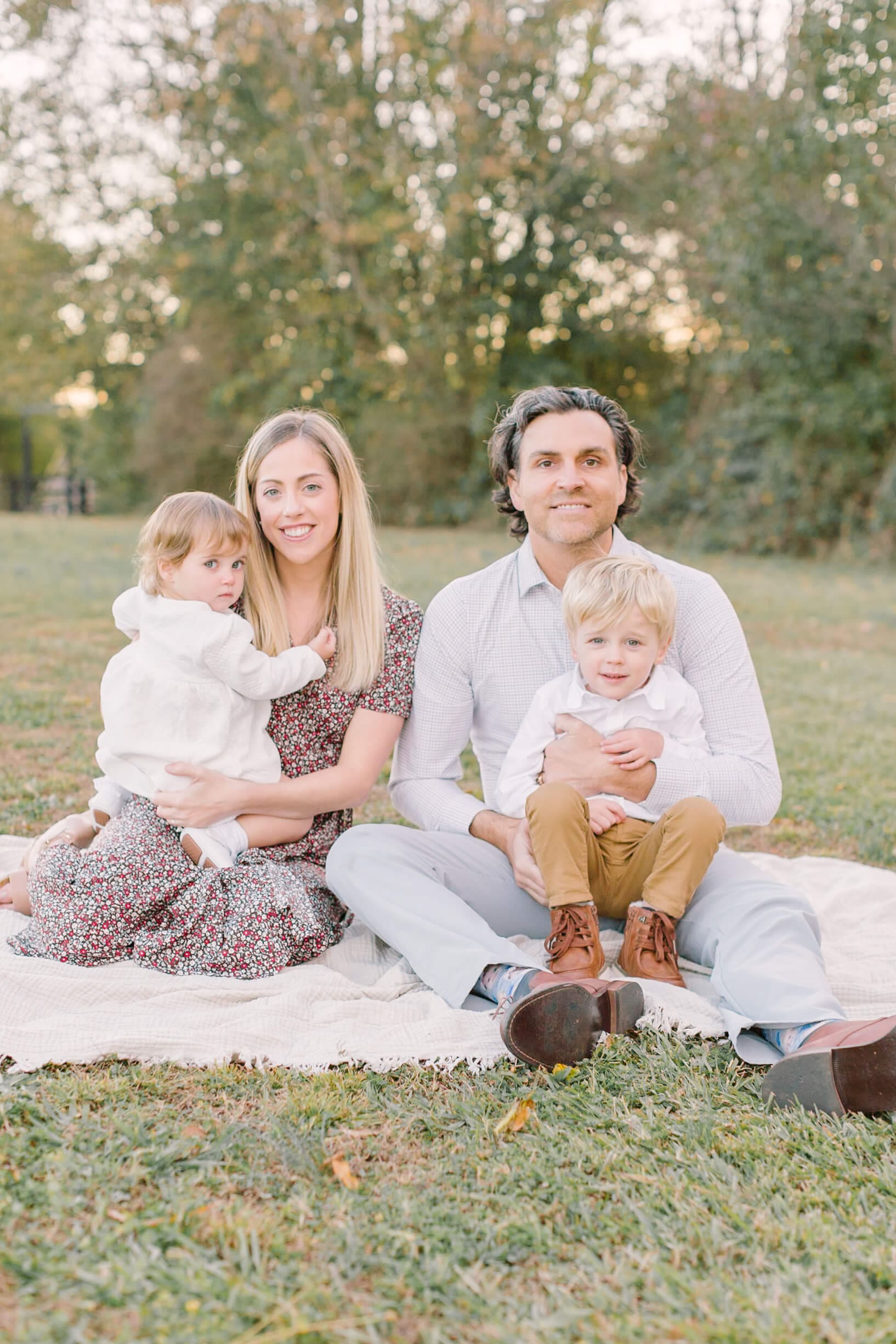 A happy mom and dad sit on a picnic blanket in a park lawn with their infant son and daughter in their laps