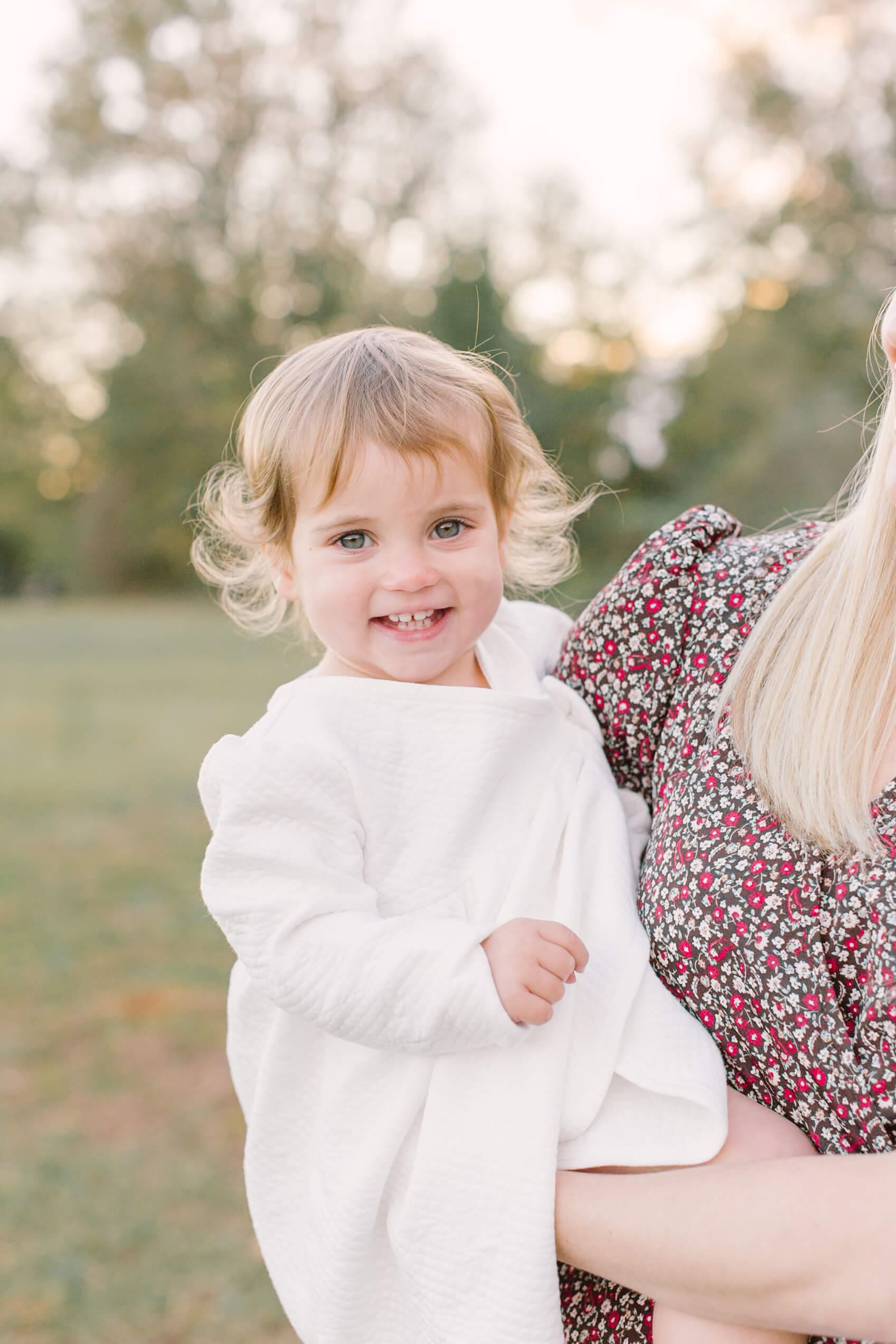 A happy toddler girl in a white dress sits in mom's arms in a park at sunset before visiting a corn maze in raleigh