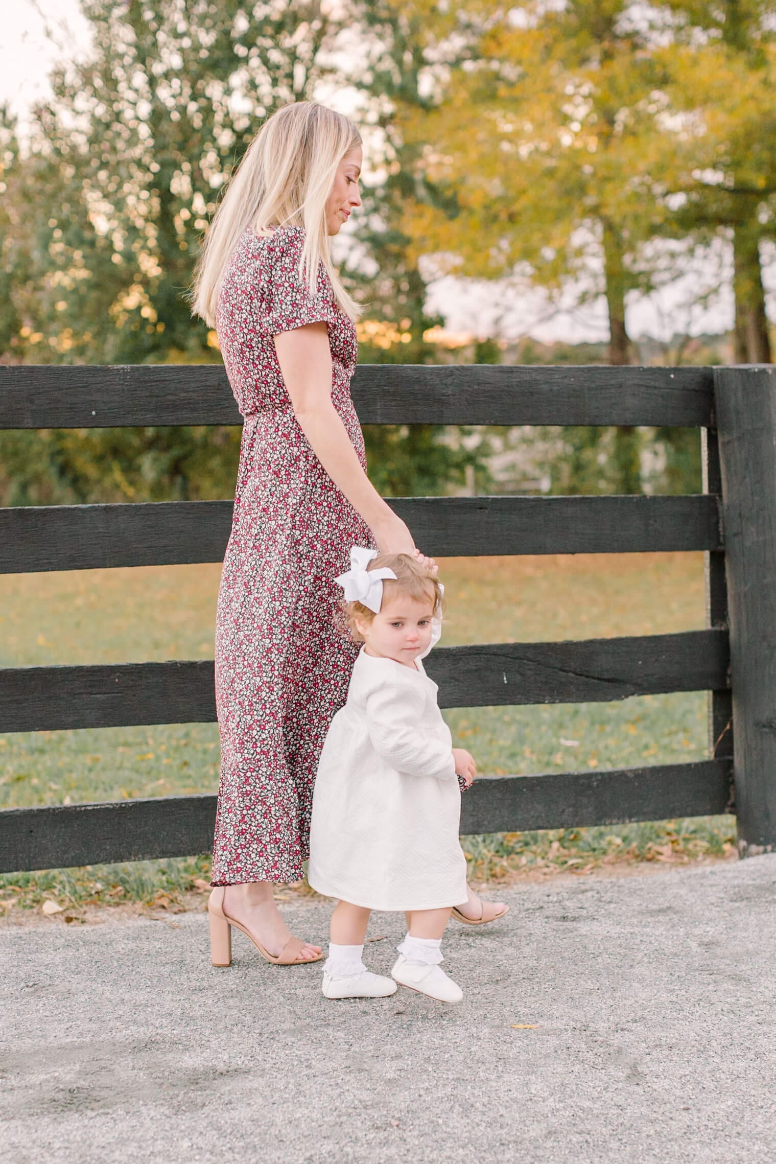 A young toddler girl in a white dress walks a park sidewalk holding mom's hand before visiting a corn maze in raleigh