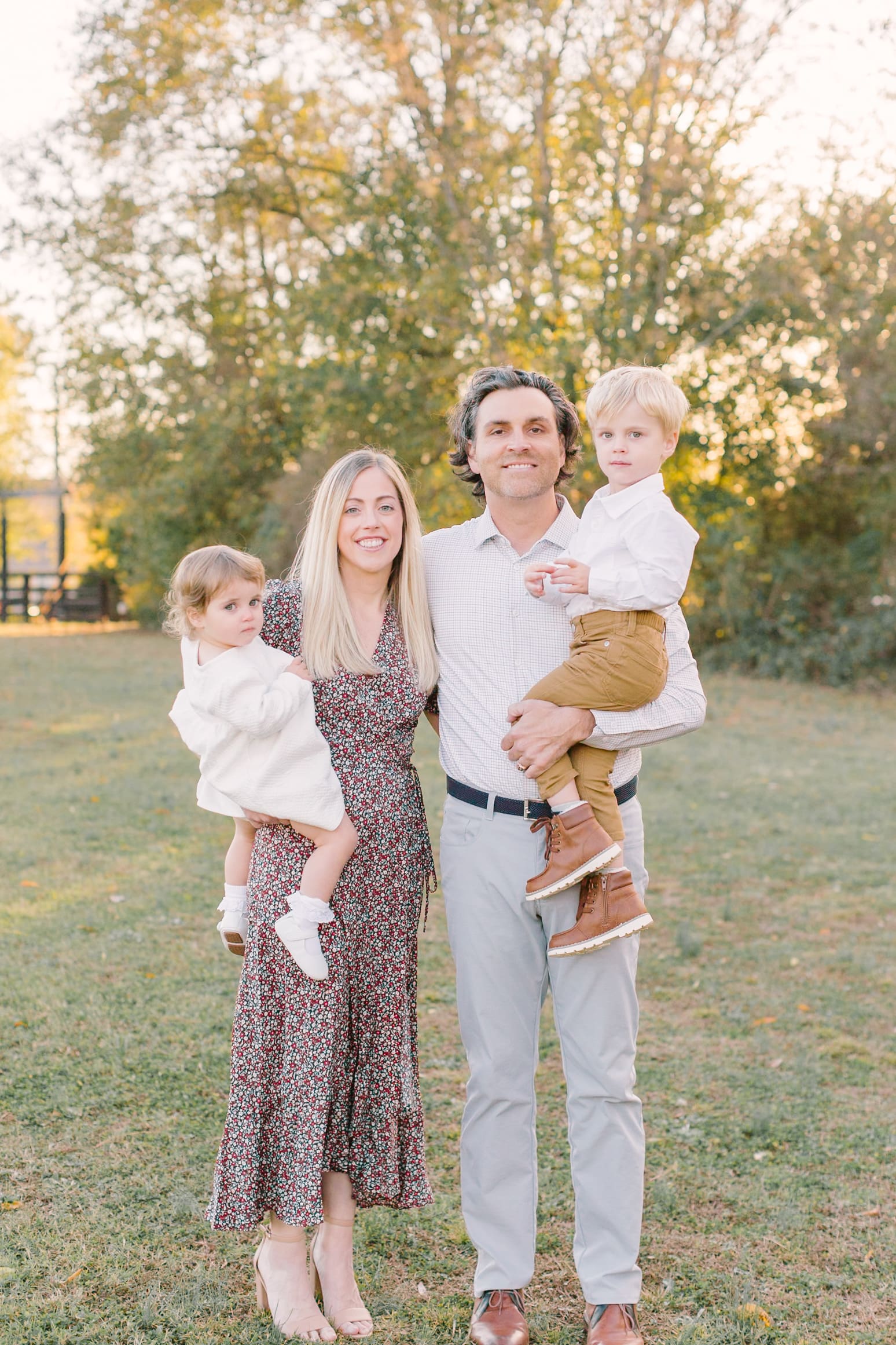 A smiling mom and dad stand in a lawn at sunset holding their toddler son and daughter on their hips