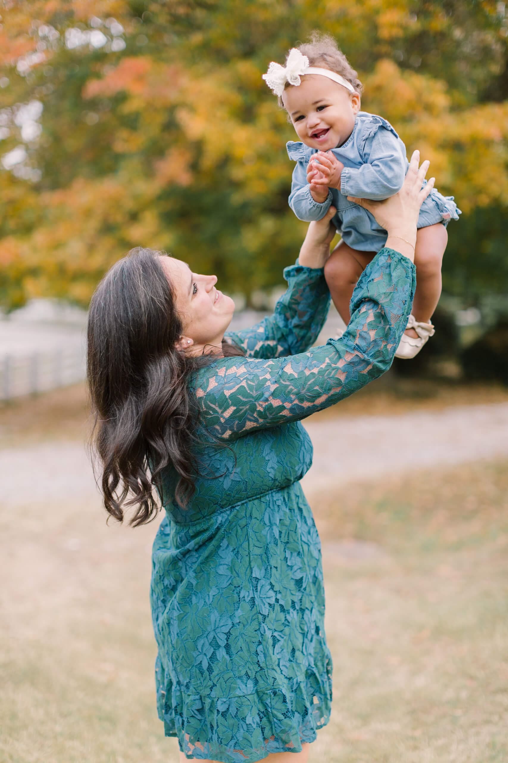 A happy infant girl in a denim dress is tossed in the air by mom in a blue dress in the lawn at sunset