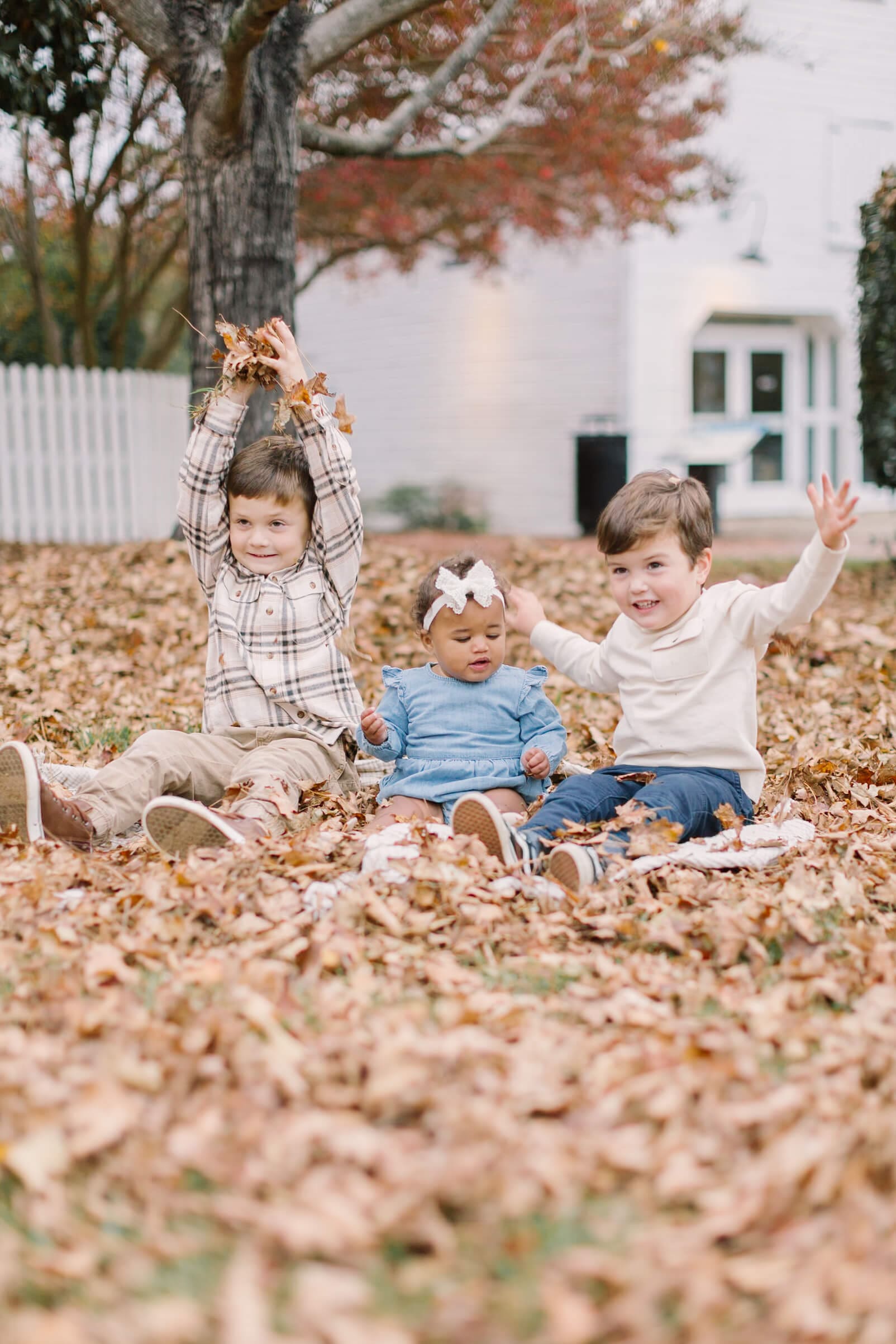 Three siblings sit and play in a leaf covered lawn during some fall activities in raleigh