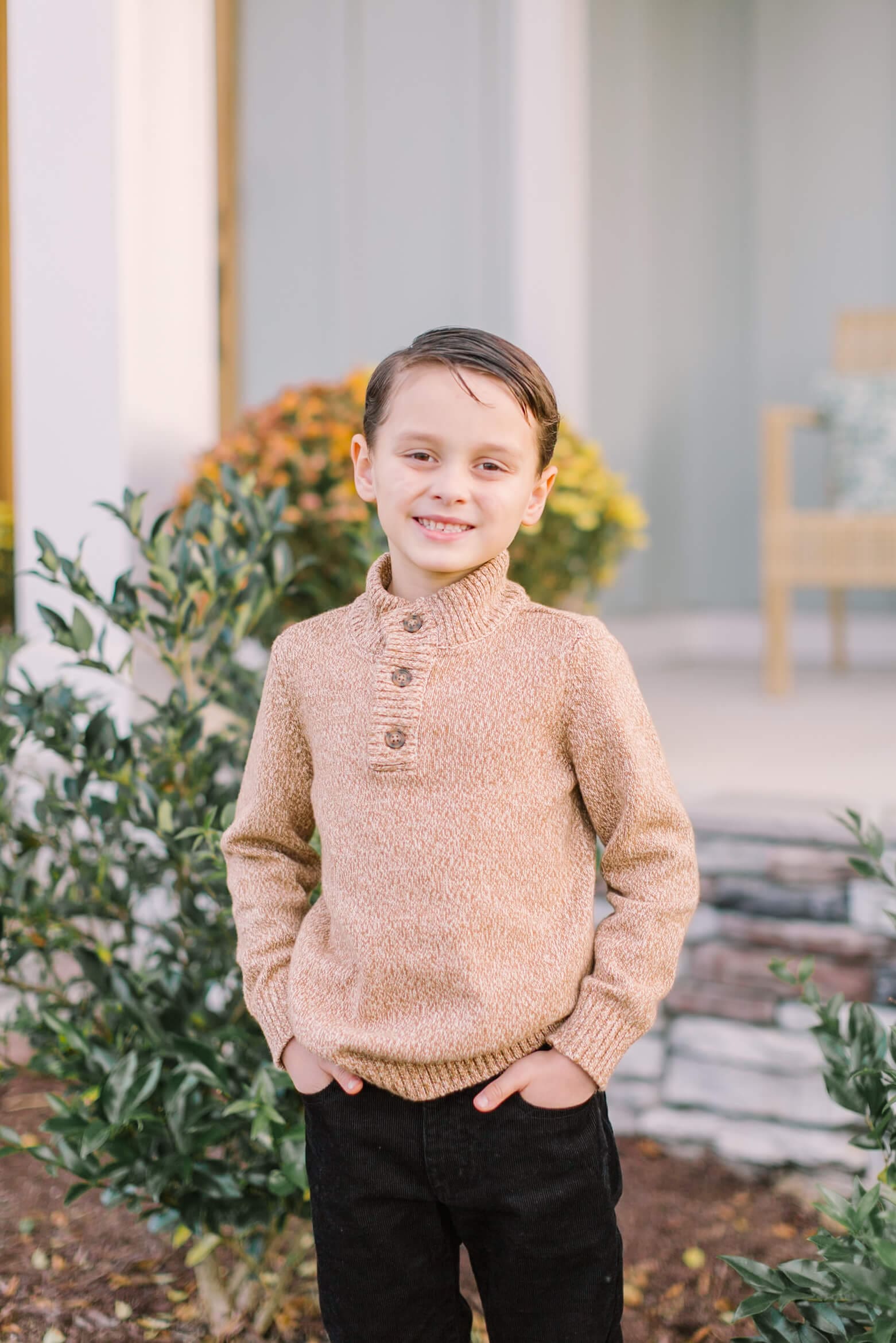 A young boy in a tan sweater smiles while standing in a garden with hands in his black jean pockets