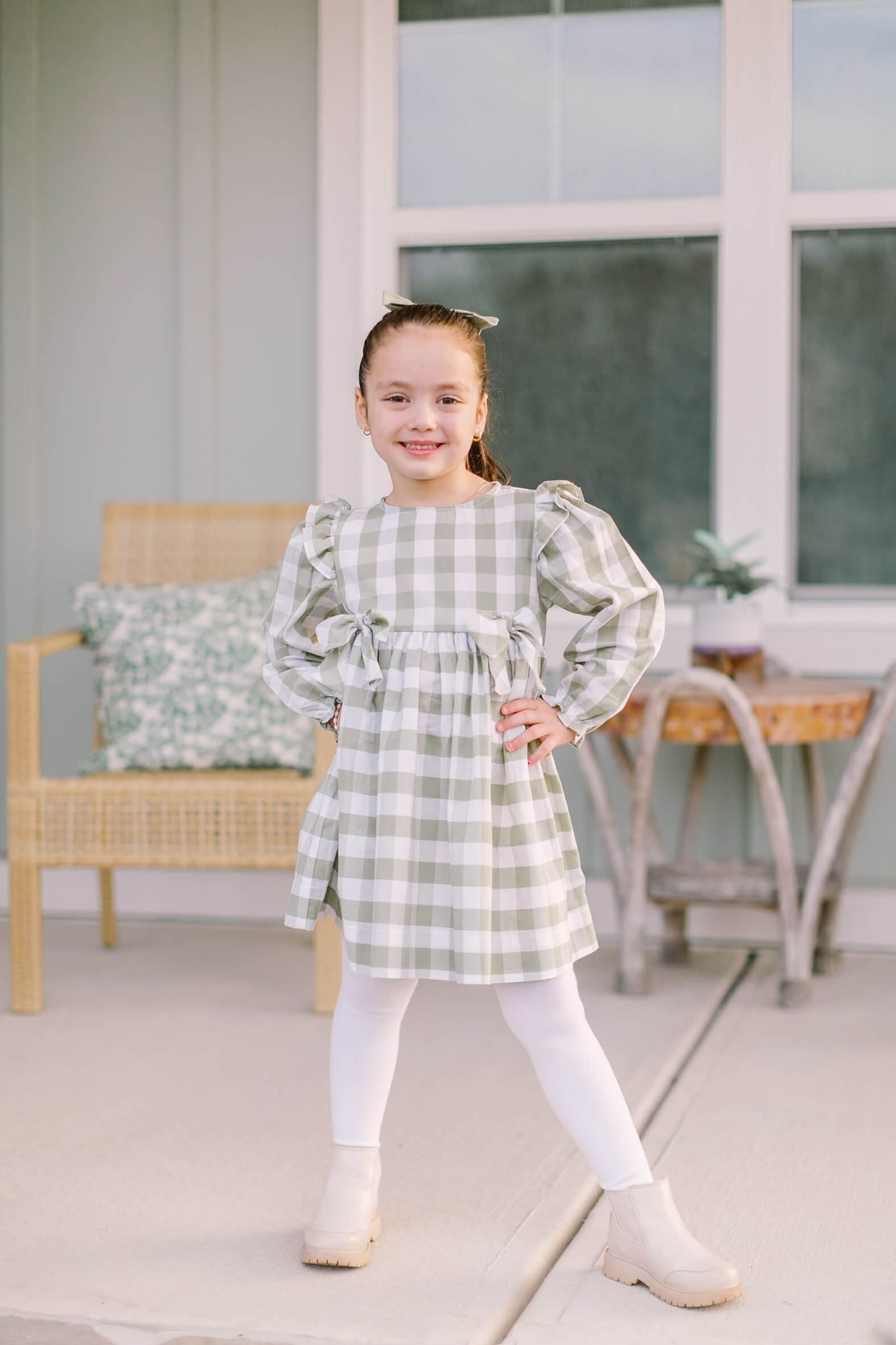 A confident young girl in a plaid dress stands with hands on her hips on a porch before visiting fall festivals in raleigh