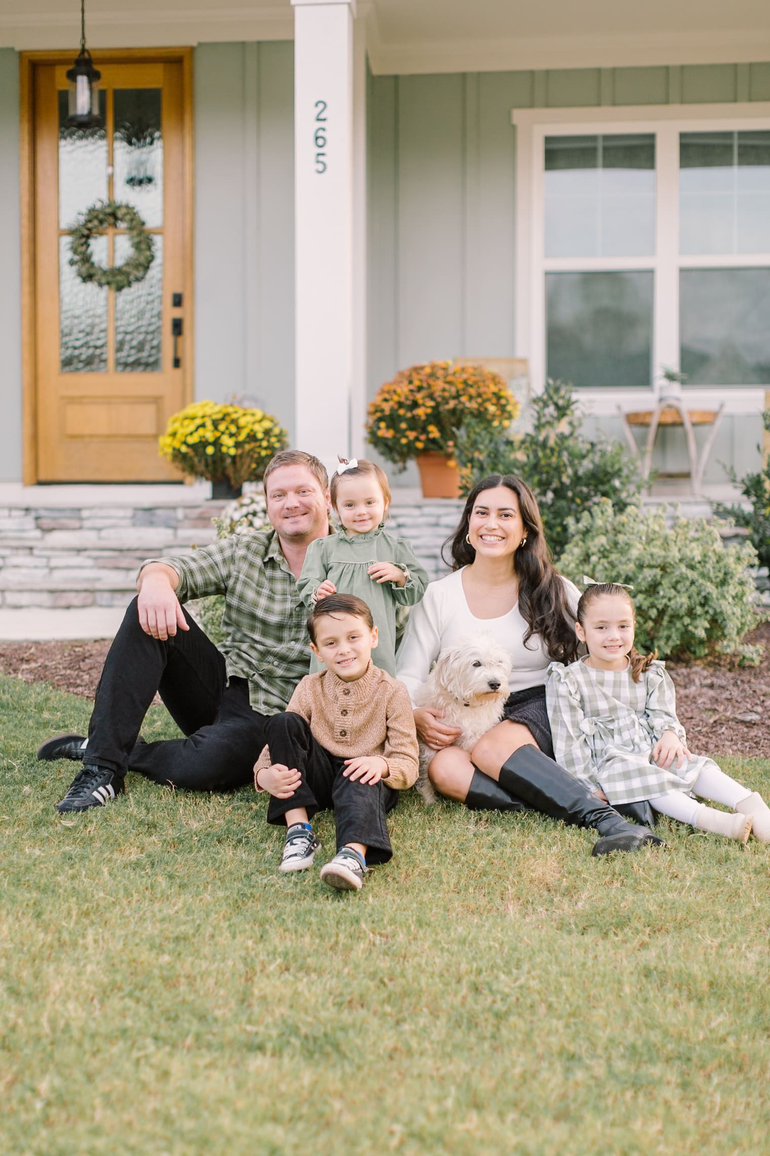 A happy family of five sit in the lawn in front of their house with a white dog in mom's lap