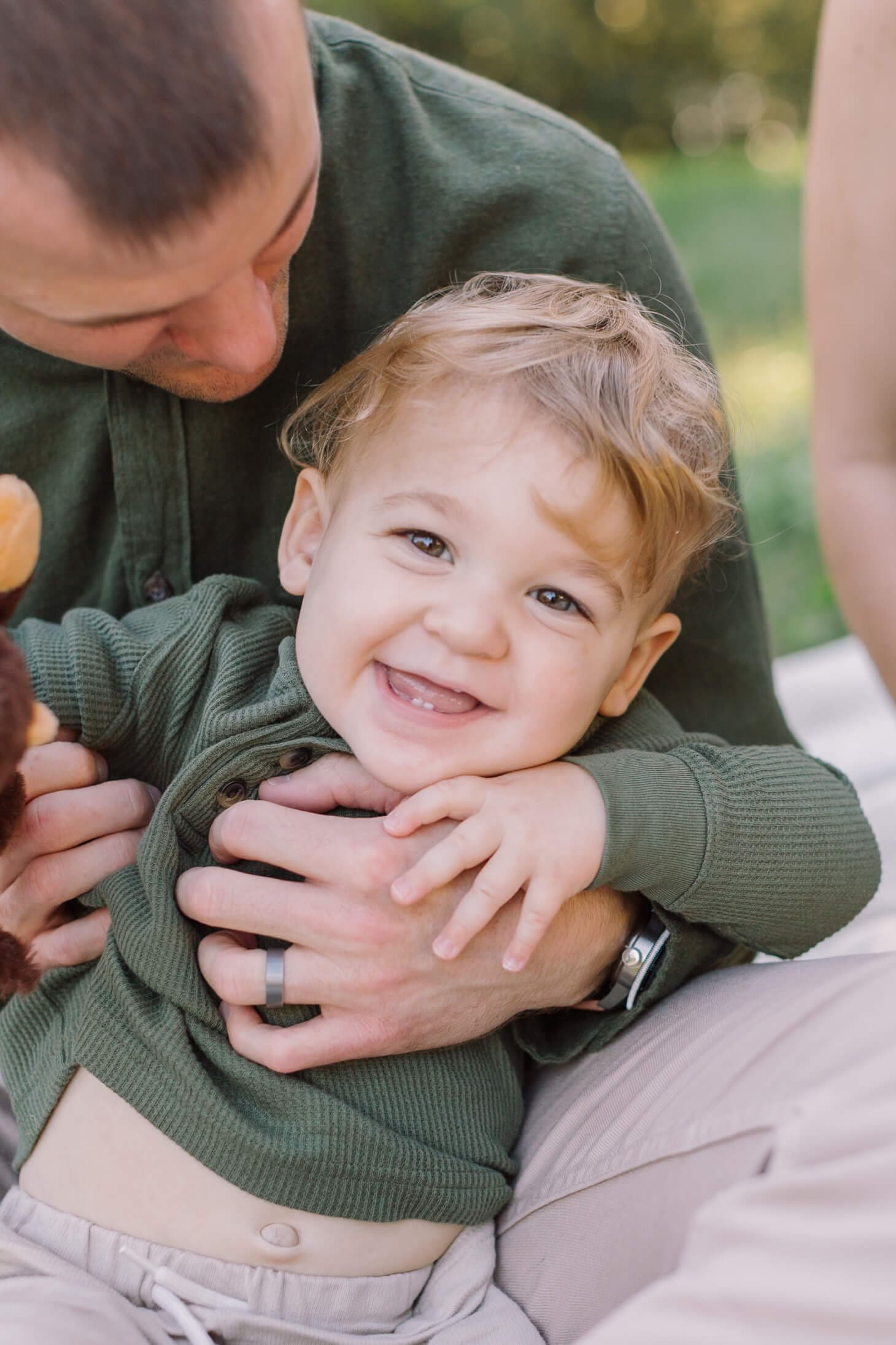 A toddler boy in a green shirt is tickled while playing in dad's lap before visiting a pumpkin patch in raleigh